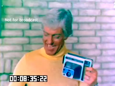 The still features a smiling man with light-colored hair, wearing a bright yellow turtleneck. He appears enthusiastic as he holds a Kodak camera, showcasing it against a backdrop of light-colored brick. The image includes a timestamp indicating a specific moment in the film. The words 'Not for broadcast' are visibly overlayed on the top of the frame. The overall tone suggests a promotional context for Kodak products from 1977.