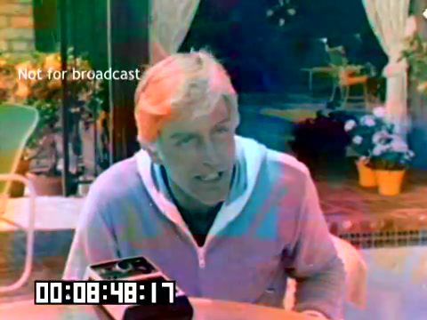 The still features a man, likely a well-known actor from the 1970s, sitting outdoors at a table with a camera positioned in front of him. He has light-colored, slightly tousled hair and is wearing a light blue zip-up jacket. The background shows a sunny, relaxed setting with plants and a pool area, suggesting a casual promotional context. The image has a slightly faded or color-altered look characteristic of older film, and a timestamp indicates its position in the overall footage. The 'Not for broadcast' text suggests that this is a restricted promotional or behind-the-scenes style content.