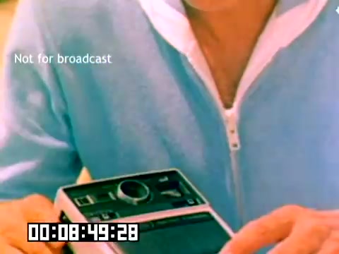 The still features a close-up of a person’s hands holding a Kodak camera, likely set against a colorful backdrop. The individual is wearing a light blue zip-up sweater. A timecode at the bottom reads '00:08:49:28' along with the text 'Not for broadcast.' The focus is on the camera, suggesting a demonstration or promotional context typical of sales films from the era. The absence of sound might limit the understanding of the full content.