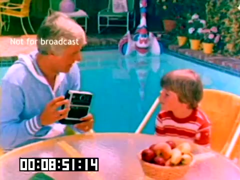 The still features two individuals by a swimming pool, likely filmed in a sunny, outdoor setting. One person, dressed in a light blue sweater, is holding a camera and engaging with a young boy, who is wearing a striped red and white shirt. There are colorful planters and a playful atmosphere, indicative of a family-friendly context. The scene is marked with timecode and a 'Not for broadcast' label, suggesting it's an archival or promotional piece, potentially highlighting Kodak's products from the era.