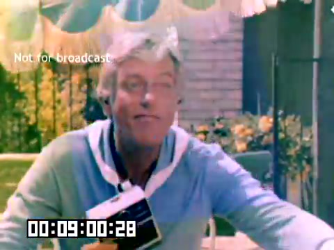 The still features a man in a light blue sweater outdoors, likely promoting Kodak products. He's holding a camera and smiling, suggesting enthusiasm for photography. The background includes a patio area with colorful decorations and blooming flowers. The image is marked with 'Not for broadcast' and has a timecode at the bottom, indicating it is a still from a sales film.