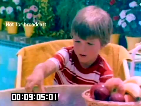 The still captures a young child sitting at a table by a swimming pool, surrounded by a vibrant setting filled with flowers and colorful decorations. The child, wearing a striped red and white shirt, appears to be intently focused on a bowl of fruit, possibly reaching for an apple. The composition is bright and cheerful, reflective of 1970s aesthetics, with the text 'Not for broadcast' overlaying the image. The overall mood suggests a leisurely, sunny day.