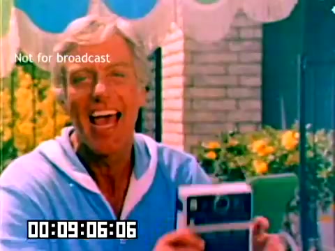 The still features a smiling man holding a Kodak camera, dressed in a light blue sweater. He appears cheerful and engaged with the audience, set against a backdrop of colorful flowers and a bright, sunny atmosphere. The image has a vibrant color palette typical of 1970s marketing materials. It includes a timecode and a watermark indicating it is not for broadcast.