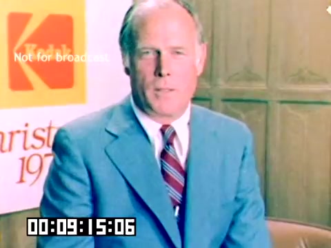 The still features a man in a blue suit and red tie sitting in front of a backdrop with the Kodak logo and the year '1977.' The image has a vintage feel typical of the 1970s sales presentations, capturing a promotional moment without sound. A timestamp in the bottom left shows 00:09:15:06, indicating a point in the film. The setting appears formal, suited for a corporate presentation.