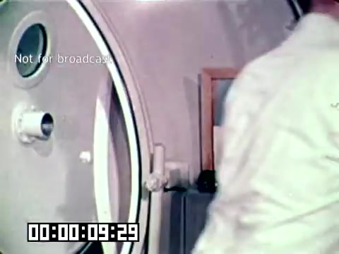 The still features a medical professional in a white coat near a hyperbaric chamber, likely illustrating the treatment for gangrene in a military context. The chamber is designed for administering oxygen therapy, emphasizing the medical advancements used to save lives. The timecode indicates it is from the early segment of the film.