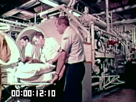 The still depicts a medical scene from the Air Force newsfilm 'Stopping gangrene - saving lives.' It shows medical personnel attending to a patient on a stretcher in a facility equipped with tubes and machinery, suggesting a focus on advanced treatments. The urgency and seriousness of the situation are evident, highlighting the importance of addressing gangrene in critical care settings.
