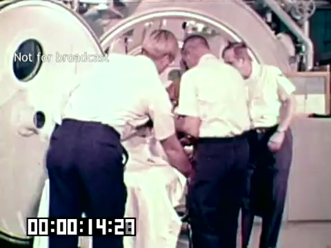The still from the Air Force newsfilm 'Stopping gangrene - saving lives' depicts a group of medical personnel working together around a patient who appears to be preparing to enter a hyperbaric chamber. The scene highlights the medical team's efforts to provide treatment, emphasizing the importance of advanced technologies in saving lives from gangrene. The atmosphere conveys urgency and collaboration in a clinical setting.