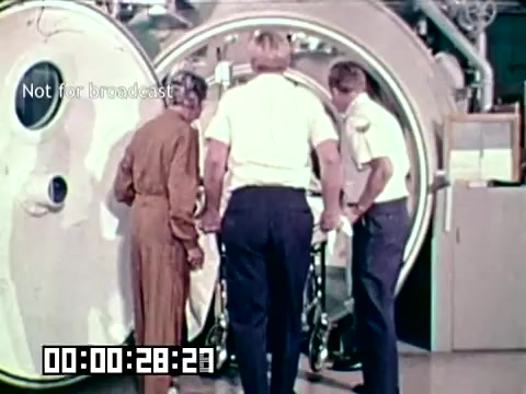 The still from the Air Force newsfilm 'Stopping gangrene - saving lives' shows three individuals examining a large hyperbaric chamber. The scene suggests a medical or research setting focused on treating conditions like gangrene, possibly involving pressure therapy. Two men in uniforms and one in a jumpsuit are engaged in a discussion near the chamber, highlighting teamwork in a medical context. The timestamp indicates an ongoing segment of the film.