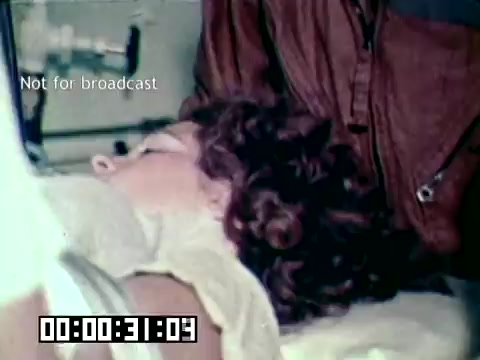 The still from the Air Force newsfilm 'Stopping gangrene - saving lives' depicts a close-up of an unconscious or injured individual in a medical setting. The patient has curly hair and appears to be receiving care, highlighting the urgency of medical intervention to prevent gangrene. The scene conveys the critical nature of military medical efforts in treating injuries.