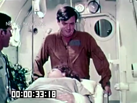 The still depicts a scene from the Air Force newsfilm 'Stopping gangrene - saving lives.' It shows a medical setting, likely within an aircraft or field hospital. A patient lies on a gurney, while two medical personnel, one wearing a flight suit and the other in surgical attire, are engaged with the patient, suggesting a focus on urgent medical care to prevent gangrene. The environment appears sterile and organized, indicative of a prioritization of medical procedures in military contexts.