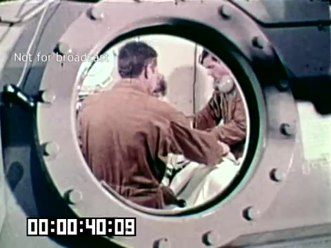The still from the Air Force newsfilm 'Stopping gangrene - saving lives' features a close-up view through a circular window of a medical environment. Inside, two medical personnel appear to be attending to a patient, emphasizing the film's focus on military medical practices aimed at preventing gangrene and saving lives. The scene conveys a sense of urgency and care in a clinical setting.