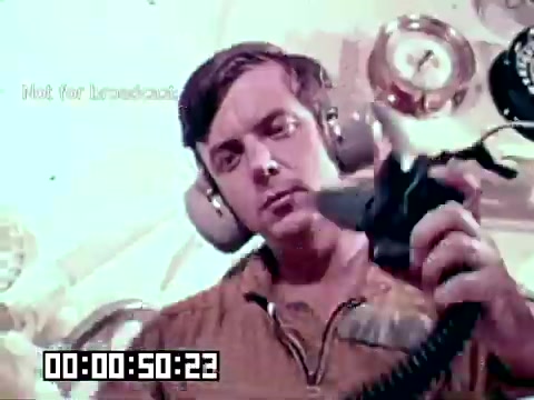 The still shows a male technician wearing headphones and holding a medical device, likely related to the treatment of gangrene. The setting appears to be within a military or aviation context, possibly a medical facility or aircraft. The focus is on the technician's concentration as he prepares to use the equipment, emphasizing the importance of medical interventions in saving lives. The image is part of an Air Force newsfilm aimed at highlighting medical advancements and procedures.