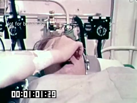 The still from the Air Force newsfilm 'Stopping gangrene - saving lives' shows a patient lying in a hospital bed. The patient appears to be undergoing treatment, with medical equipment visible in the background. An IV line is connected to the patient's arm, indicating they are receiving care. The scene likely emphasizes the importance of medical interventions in preventing gangrene and saving lives. The timestamp at the bottom suggests this is a frame captured from a specific moment in the footage.