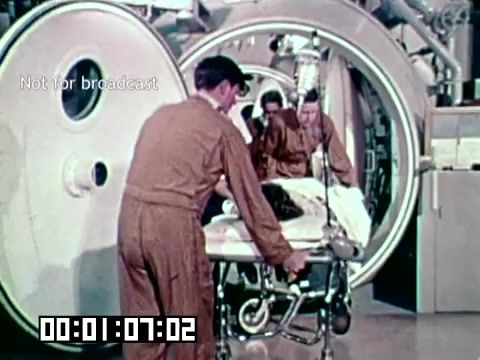 The still depicts a medical scene from the Air Force newsfilm 'Stopping gangrene - saving lives.' It shows medical personnel attending to a patient on a gurney, likely in a high-tech medical facility. The focus seems to be on the application of specialized treatment to prevent gangrene. The environment includes large, circular medical devices, suggesting a technological approach to patient care. The overall tone conveys urgency and the importance of medical intervention in life-saving situations.