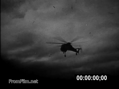 The 16mm still from 'Your Man In Vietnam' depicts a CH-54 helicopter flying against a cloudy sky, capturing the dramatic atmosphere of military operations during the Vietnam War era. The image emphasizes the role of U.S. Army flight engineers like Specialist 6 James Daly, highlighting the challenges faced by service members in a wartime environment. The monochrome aesthetic reflects the historical nature of the footage, giving a glimpse into the realities of army life during this period.