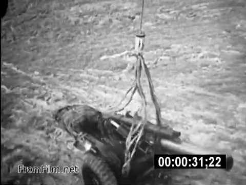The still depicts a CH-54 helicopter engaged in a military operation, showcasing the complexity of logistics during the Vietnam War. In the image, a piece of artillery is suspended from a cable, demonstrating the aircraft's capability to transport heavy equipment in challenging terrains. This highlights the critical role of flight engineers like Specialist 6 James Daly in supporting ground operations. The footage serves as a historical reminder of the everyday realities faced by U.S. Army personnel during the conflict.