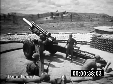 The still depicts a scene from a Vietnam War-era public service announcement featuring U.S. Army personnel operating a large artillery piece, likely a howitzer, set against a backdrop of a desolate landscape. Soldiers are engaged in preparing for or conducting a firing operation, illustrating the daily responsibilities and challenges faced by military personnel during the conflict. The image captures the gritty reality of Army life, emphasizing teamwork and the stark environment of the Vietnam War.