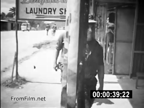 The still from 'Your Man In Vietnam' depicts a scene from the Vietnam War era featuring Specialist 6 James Daly, a CH-54 flight engineer. The image shows Daly walking past a laundry shop, highlighting everyday life for U.S. Army service members in Vietnam. The surrounding environment reflects the military presence and local culture during the period, capturing a moment of routine amidst the backdrop of war. This PSA aims to present the relatable experiences of soldiers, emphasizing their daily challenges and contributions.