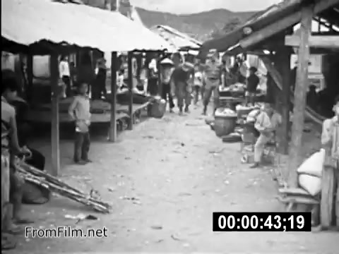 The 16mm still from 'Your Man In Vietnam' depicts a bustling scene set in a village during the Vietnam War. In the foreground, it shows a dirt path lined with makeshift stalls and vendors, highlighting the local economy. U.S. Army personnel are seen interacting with local residents, suggesting themes of community engagement and daily life. The setting is reflective of the everyday realities confrontational for soldiers like Specialist 6 James Daly, emphasizing the human connections and challenges faced during their service. The black-and-white footage captures a historical moment, providing insight into military life and the local culture during the era.