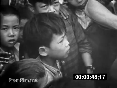 This 16mm still from the public service announcement 'Your Man In Vietnam' depicts a group of Vietnamese children closely observing something off-camera, likely related to U.S. military activities. A young boy in the foreground stands out with a contemplative expression, embodying a moment of curiosity or concern amid the backdrop of the Vietnam War. The image captures the intersection of military presence and civilian life during this historical period, emphasizing the personal stories affected by the conflict.