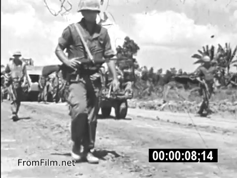 The still from 'Your Man In Vietnam' depicts U.S. Army soldiers on a dirt road, conveying the rugged and challenging environment of the Vietnam War. In the foreground, a soldier in a helmet and military fatigues marches purposefully, likely emphasizing the realities of military life. In the background, other soldiers can be seen, along with military vehicles, suggesting ongoing operations. The stark imagery highlights the historical context and experiences of service members during the Vietnam era.