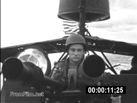 The still captures a moment from the public service announcements featuring Specialist 6 James Daly, a flight engineer aboard a CH-54 helicopter during the Vietnam War. The scene shows him seated in the cockpit, focused on the controls while armed with visible machine guns. His helmet and serious expression emphasize the tension of military life in the era. The backdrop suggests a clouded sky, hinting at the challenges faced by service members in Vietnam. This footage highlights both the operational aspects of Army life and the personal experiences of those serving during the conflict.