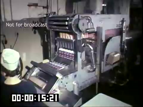 The still shows a vintage processing machine in a food manufacturing facility, specifically related to the production of frozen novelties. It features a worker in a cap, operating the machine, which has a series of colorful products in view. The setting appears industrial, with various controls visible on the equipment. The image reflects the production techniques of the 1950s-1960s, highlighting the technology of that era. The frame has a timestamp indicating it's part of a larger video and has a “Not for broadcast” label.