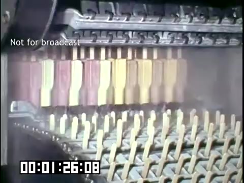 The still from 'DCA Food Industries - Making Frozen Novelties' features a close-up of a production line that appears to be filling molds with colorful mixtures for frozen treats. The scene captures several rows of molds in various pastel shades, indicating an efficient assembly process typical of the 1950s-1960s, showcasing the industrial production of frozen novelties. The image has a nostalgic aesthetic, characteristic of Ektachrome film stock from that era.