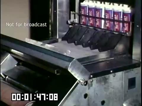 The still features a mechanical process involved in the production of frozen novelties at DCA Food Industries. It showcases a metal processing machine with empty cartons positioned to be filled. The machine is part of a larger assembly line, indicative of the industrial practices of the 1950s-1960s. The image captures the efficiency and technological advancements in food manufacturing during that era. The 'Not for broadcast' watermark implies this footage was intended for internal or educational purposes rather than public release.