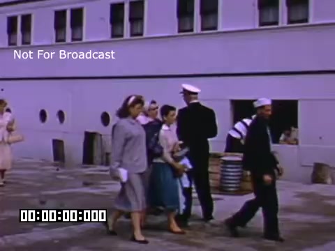 The still from 'In and Around Battle Creek Michigan' features a scene with several individuals walking on a paved area. The setting appears to be an outdoor location, likely near a waterfront or dockside, given the presence of a uniformed sailor and various structures in the background. The individuals are dressed in mid-20th century attire, typical of the 1950s, with some women wearing dresses and the man in a navy uniform. The colors are vibrant, indicative of Kodachrome film, which captures bright, saturated hues. The scene conveys a sense of casual interaction among the group, reflecting the social atmosphere of the time.