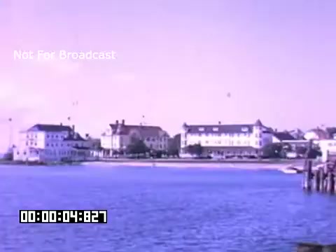 The still from 'In And Around Battle Creek Michigan' showcases a serene water scene, likely featuring a lakeside or waterfront view. The image captures a collection of buildings along the shore, characterized by their traditional architecture. The colors are soft and pastel, typical of Kodachrome film, lending a nostalgic vibe. The presence of gentle water indicates a calm atmosphere, while the structures suggest a community or resort area. The timestamp indicates this is an excerpt from a larger film. Overall, it embodies a tranquil snapshot of mid-20th century American life.