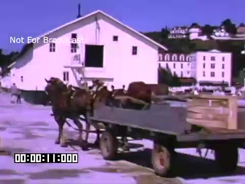 The still from 'In And Around Battle Creek Michigan' depicts a horse-drawn wagon unloading crates near a large building, likely part of a production or agricultural setting. The scene is captured in vibrant Kodachrome color, reflecting a historical context. The architecture in the background features a distinctive roofline, suggesting a commercial or warehouse purpose. The overall atmosphere conveys a lively, industrious environment from the period.
