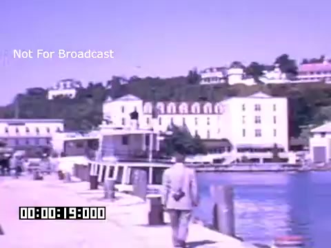 The still features a picturesque scene near a waterfront in Battle Creek, Michigan, showcasing a series of buildings on a hillside. A man dressed in a light-colored suit walks along a dock, with boats moored nearby. The color palette indicates a vintage aesthetic, typical of Kodachrome footage, with soft, vibrant hues. The overall composition suggests a serene and inviting atmosphere, highlighting the charm of the area.