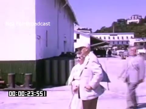 The still from 'In And Around Battle Creek Michigan' features a scene with several people outdoors near a large building. The setting appears to be a sunny, warm day, with bright colors typical of Kodachrome film. In the foreground, two individuals—one older man in a light suit and a woman in a white outfit—are seen standing together. They appear to be engaging in conversation or observing their surroundings, while other figures and buildings are visible in the background, suggesting an active outdoor environment. The scene captures a moment of everyday life, reflecting the culture and atmosphere of the time in Battle Creek, Michigan.