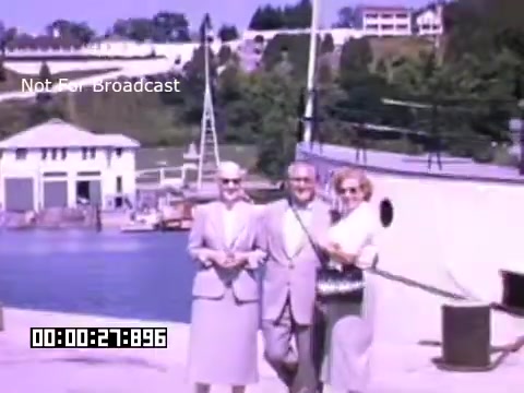 The still depicts three individuals standing in front of a body of water with a dock and a building in the background. The scene has a nostalgic, vintage feel, typical of 16mm Kodachrome footage. The individuals appear dressed in smart casual attire, suggesting a leisure outing. Lush, green trees and a clear sky provide a serene backdrop. The text 'Not For Broadcast' is visible, indicating the footage is not intended for public viewing. The overall mood conveys a sense of camaraderie and enjoyment in the setting of Battle Creek, Michigan.