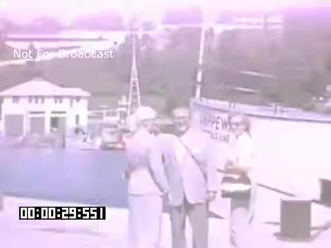 The still from 'In And Around Battle Creek Michigan' features a group of people outdoors, likely in a recreational setting. They are dressed in mid-20th century attire, suggesting a casual gathering or event. The background includes structures typical of the era and hints at a community atmosphere. The colors are slightly faded, characteristic of Kodachrome footage, giving the image a nostalgic feel. The timestamp indicates it's a brief moment in the video.