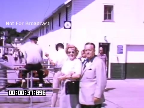 The still from 'In And Around Battle Creek Michigan' features a vibrant outdoor scene captured in Kodachrome, indicative of mid-20th century Americana. In the foreground, a couple poses together, dressed in formal attire, exhibiting a sense of camaraderie and enjoyment. The background reveals a bustling environment, with people engaging in various activities and a building suggesting a community or recreational space. Soft pastel colors characterize the image, evoking a nostalgic vibe typical of the era. The inclusion of a timestamp and the note 'Not For Broadcast' adds a layer of context regarding its intended use.