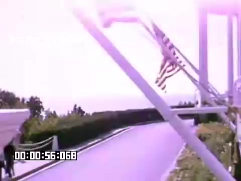 The still from 'In And Around Battle Creek Michigan' features a bright scene with a prominent American flag waving in the foreground. The composition suggests a focus on the landscape surrounding a road or path, which is bordered by greenery. The overall color palette reflects the vibrant hues typical of Kodachrome film, giving it a nostalgic feel. A timestamp indicating 56 seconds into the footage is visible at the bottom. The atmosphere appears idyllic, potentially capturing a moment of serene outdoor life in the area.