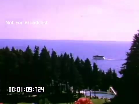 The still from 'In And Around Battle Creek Michigan' features a picturesque view of a lake scene. In the foreground, a lush area with trees can be seen, while the background showcases a serene blue lake. A boat is moving across the water, adding a dynamic element to the tranquil landscape. The vibrant colors suggest a warm, sunny day, typical of Kodachrome footage. The timestamp indicates a moment captured in the film, while a label indicates it is not for broadcast.