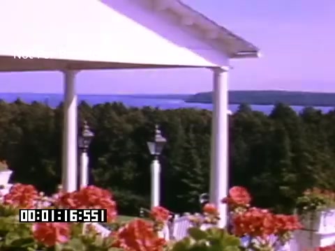 The 16mm still from 'In And Around Battle Creek Michigan' features a picturesque view, likely taken during a bright day. In the foreground, vibrant red flowers bloom in planters, adding a lively touch to the scene. Behind them, a white porch or building structure is visible, framed by columns. Beyond the architecture, lush greenery and a body of water can be seen in the background, suggesting a serene natural landscape. The shot captures a peaceful ambiance, indicative of a scenic location in Michigan.