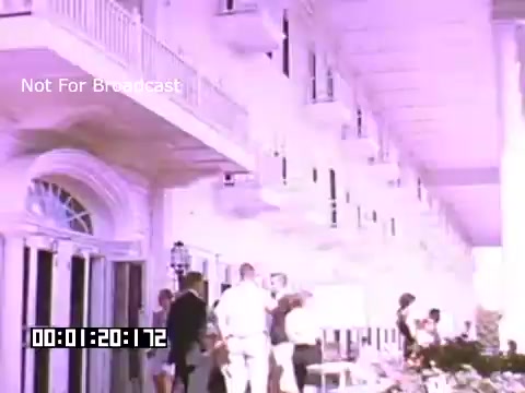 The still from 'In And Around Battle Creek Michigan' features a bustling scene outside a building, likely a hotel or resort. People are gathered in various social interactions, dressed in mid-20th century attire. The architecture has an elegant style, with balconies and large windows. The color palette is soft, typical of Kodachrome footage, hinting at a warm atmosphere. The scene encapsulates a lively social gathering in a classic American setting.