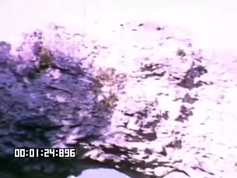 The still features a rocky terrain, possibly a cliff or a natural formation, with patches of vegetation visible among the rocks. The color palette is soft, likely due to the Kodachrome processing, giving it a nostalgic and vintage appearance. The timestamp at the bottom indicates the exact position in the footage. The background is slightly blurred, focusing attention on the rock formation.