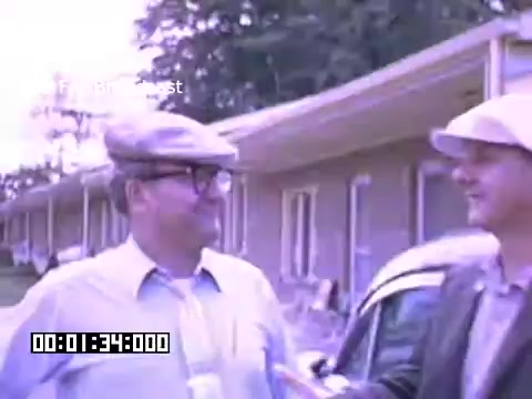 The still features two men conversing outside a row of buildings, likely a residential or commercial area in Battle Creek, Michigan. One man is wearing a flat cap and glasses, while the other has a light-colored cap. The colors are vibrant and indicative of Kodachrome film, capturing a casual, possibly nostalgic moment in a mid-20th century setting. The timestamp suggests this is an excerpt from a longer film.