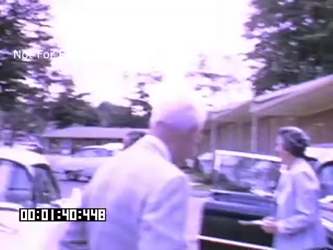 The still frame from 'In And Around Battle Creek Michigan' features a scene with two individuals—one man in a light suit and a woman in a gray outfit—interacting near parked cars. The background includes trees and what appears to be a building, indicating a suburban or community setting. The colors are slightly washed out, characteristic of Kodachrome footage, giving it a nostalgic and vintage feel. The timecode in the corner suggests a specific moment in the film.