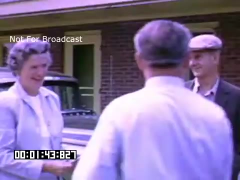 The still captures a moment from 'In And Around Battle Creek Michigan,' featuring three individuals engaging in conversation outdoors. A woman in a light-colored outfit is extending her hand, possibly greeting the two men, who appear to be wearing hats and casual attire. The background suggests a residential setting, hinting at a warm, friendly interaction. The vibrant colors from the Kodachrome footage evoke a nostalgic atmosphere typical of mid-20th century American life.