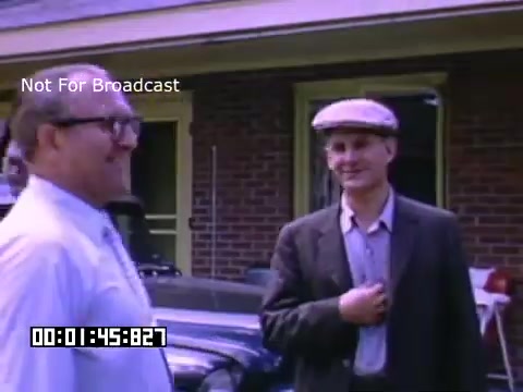 The still from 'In And Around Battle Creek Michigan' shows two men standing in front of a brick house, smiling and engaging with each other. One man, wearing glasses and a white shirt, appears to be older, while the other, in a suit and flat cap, looks slightly younger. There are vintage cars parked nearby, and the overall scene reflects a casual, conversational moment typical of mid-20th century American life. The colors suggest the use of Kodachrome film, enhancing the nostalgic feel of the image.