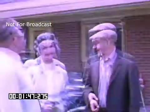 The still from 'In And Around Battle Creek Michigan' showcases a group of people in a casual outdoor setting. They appear to be engaged in conversation, with one person holding a cigarette. The colors are vivid, characteristic of Kodachrome footage, reflecting a warm, nostalgic vibe. The background suggests a residential area, with a brick house visible, contributing to the intimate, community-oriented atmosphere of the scene.