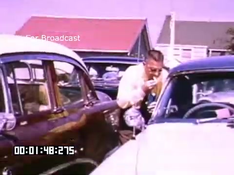 The still from 'In And Around Battle Creek Michigan' features a man interacting with a car, possibly checking something under the hood or adjusting a part. The scene is set in a vibrant environment with colorful Kodachrome hues, typical of mid-20th century American life. Nearby cars reflect the style of the era, showcasing vintage designs. The background hints at a suburban setting, possibly with houses or structures in soft pastel colors. Overall, the image captures a nostalgic moment in automotive culture from that time.