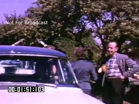 The still shows a vintage scene from 'In And Around Battle Creek Michigan,' featuring several people interacting near a classic car. The individuals appear to be enjoying a casual moment, possibly outdoors in a park or residential area, surrounded by trees. The color palette, typical of Kodachrome film, gives the image a warm, nostalgic feel. The atmosphere seems relaxed and friendly, reflecting a social gathering. The timestamp and 'Not For Broadcast' label indicate that this still is intended for archival or private viewing purposes.