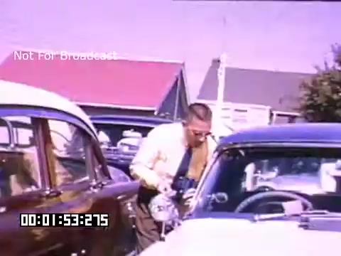 The still from 'In And Around Battle Creek Michigan' captures a moment in a vintage scene featuring a man wearing a light-colored shirt, presumably examining or interacting with a car. The setting appears to be outdoors, evident from the bright colors of the vehicles and the background, including buildings with a pinkish hue. The image conveys a sense of nostalgia, characteristic of the Kodachrome footage era, emphasizing daily life and automotive culture of the time.